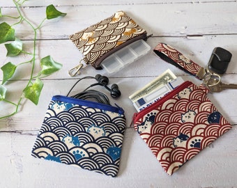 May include: Three small zippered pouches with a cat and wave pattern. The pouches are in blue, red, and brown. The pouches are on a white wooden surface.