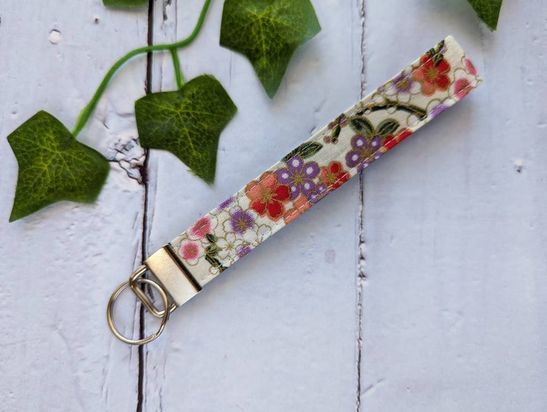 May include: A white fabric key chain with a floral pattern in shades of pink, red, and purple. The key chain has a silver metal ring.
