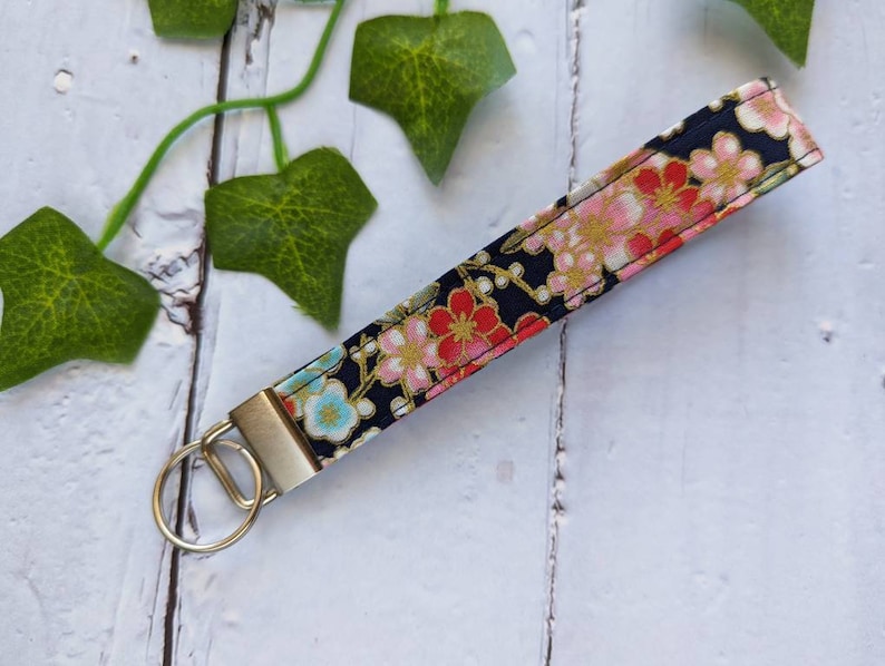 May include: A black fabric keychain with a floral pattern in pink, red, and gold. The keychain has a silver metal ring.