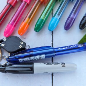 May include: A collection of colorful pens, including red, pink, orange, green, blue, purple, and black. A blue Mini RSVP pen and a black Sharpie marker are also visible, along with a small black keychain light.