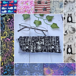 May include: A black and white glasses case with a science theme. The case has a flap that folds over to protect the glasses. The case is made of fabric and has a zipper closure. The case is sitting on a white surface with green ivy.