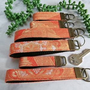 May include: Five orange and white patterned keychains with black backing and gold key rings. The keychains are arranged in a stack with the top keychain having a gold key ring attached to a silver key.