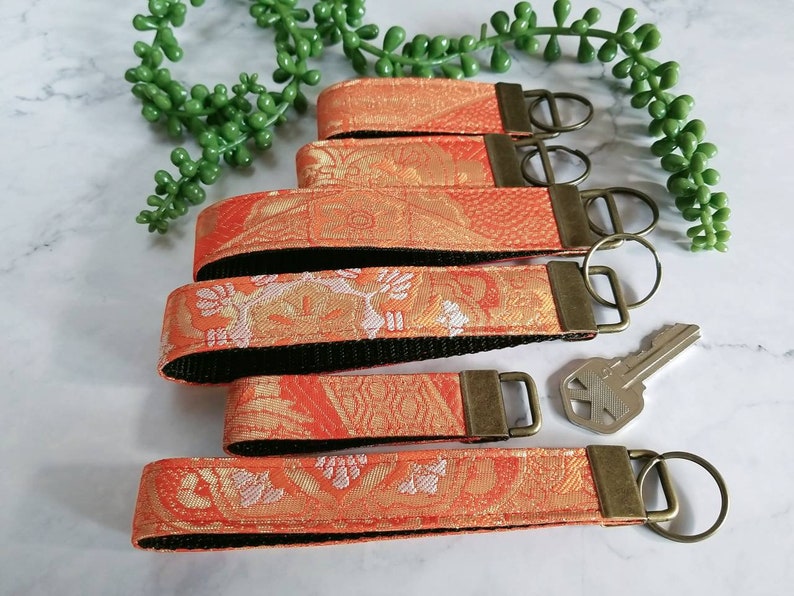 May include: Five orange and white patterned keychains with black backing and gold key rings. The keychains are arranged in a stack with the top keychain having a gold key ring attached to a silver key.