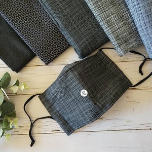 May include: A black face mask with a gray and white patterned design. The mask has a black elastic band and a white label that says "XL".