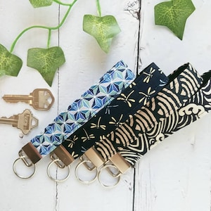 May include: Three keychains with different fabric patterns. The keychain on the left has a blue and white geometric pattern. The middle keychain has a black fabric with a gold dragonfly pattern. The keychain on the right has a black and gold fabric with a wave pattern.