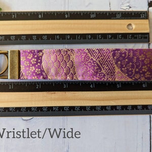 May include: A purple and gold fabric wristlet keychain with a gold metal key ring. The fabric has a traditional Japanese pattern. The wristlet measures approximately 4 inches long.