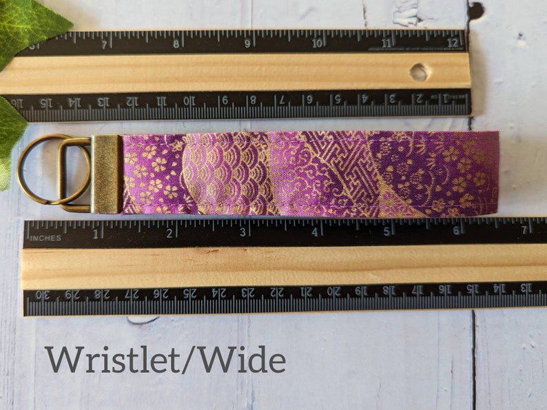 May include: A purple and gold fabric wristlet keychain with a gold metal key ring. The fabric has a traditional Japanese pattern. The wristlet measures approximately 4 inches long.