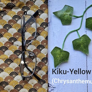 May include: A pair of black framed glasses on a yellow and brown fabric with a fan-shaped pattern. The fabric is next to a light blue surface with a green vine and the text "Kiku-Yellow (Chrysanthemum)".