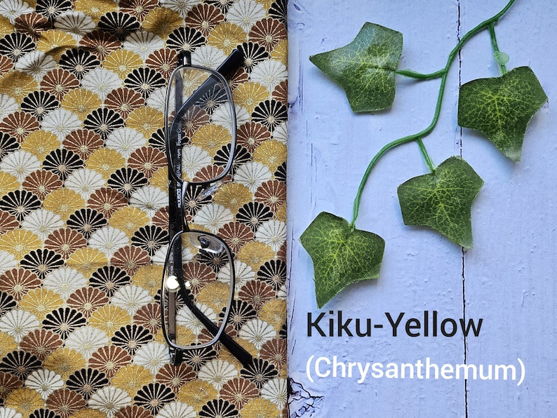 May include: A pair of black framed glasses on a yellow and brown fabric with a fan-shaped pattern. The fabric is next to a light blue surface with a green vine and the text "Kiku-Yellow (Chrysanthemum)".