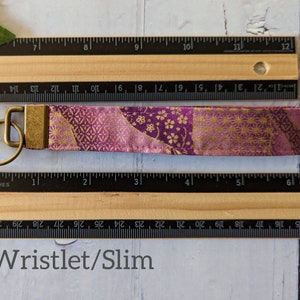 May include: A purple and gold fabric wristlet keychain with a gold metal key ring. The wristlet is approximately 3 inches long. Wristlet/Slim.