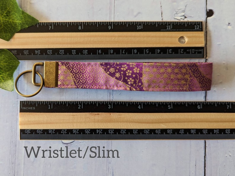 May include: A purple and gold fabric wristlet keychain with a gold metal key ring. The wristlet is approximately 3 inches long. Wristlet/Slim.