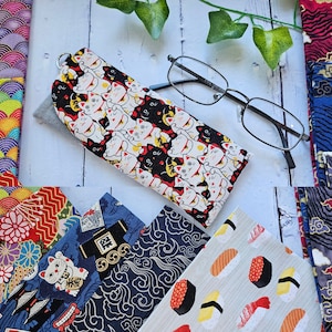 May include: A white and black cat print fabric glasses case with a silver zipper. The case is lying on a white wooden surface with various patterned fabrics in the background.