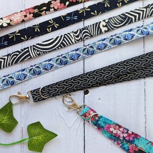 May include: Five different patterned fabric lanyards with gold metal clasps. The lanyards are arranged in a row, with the floral patterned lanyard at the top and the blue and white geometric patterned lanyard at the bottom. The lanyards are all about 1 inch wide.