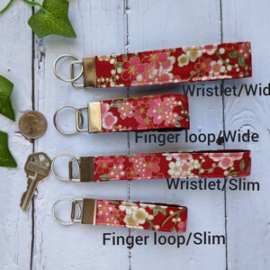 May include: Four red fabric keychains with a floral pattern. The keychains are available in two styles: wristlet and finger loop, and two widths: wide and slim. The text "Wristlet/Wide", "Finger loop/Wide", "Wristlet/Slim", and "Finger loop/Slim" is printed below each keychain.