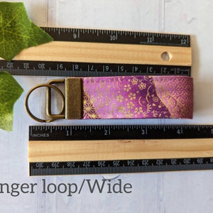 May include: A purple and gold fabric keychain with a brass key ring. The fabric has a floral pattern and is approximately 1 inch wide.