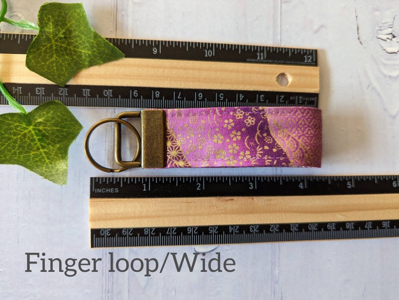 May include: A purple and gold fabric keychain with a brass key ring. The fabric has a floral pattern and is approximately 1 inch wide.