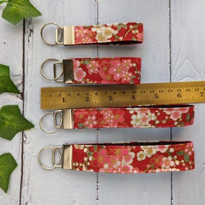 May include: Four red fabric keychains with a floral pattern and gold accents. The keychains are different lengths and are measured with a ruler in inches.