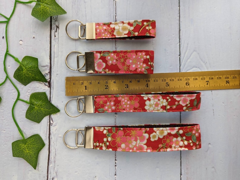 May include: Four red fabric keychains with a floral pattern and gold accents. The keychains are different lengths and are measured with a ruler in inches.