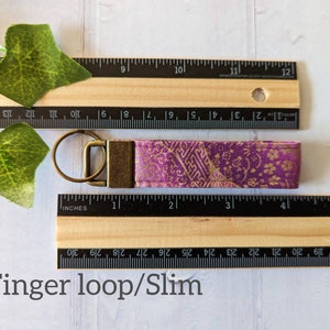 May include: A purple and gold fabric key chain with a gold metal key ring. The key chain is approximately 4 inches long. The fabric has a floral pattern.