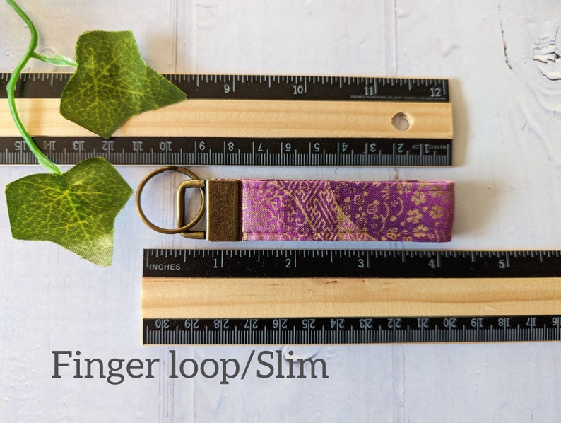 May include: A purple and gold fabric key chain with a gold metal key ring. The key chain is approximately 4 inches long. The fabric has a floral pattern.