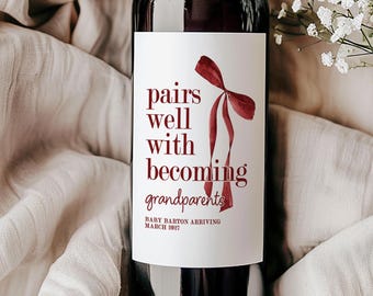 May include: A dark glass wine bottle with a white label featuring a red ribbon graphic and the text: pairs well with becoming grandparents, BABY BARTON ARRIVING MARCH 2017. The bottle is surrounded by soft, light-colored fabric and white flowers.