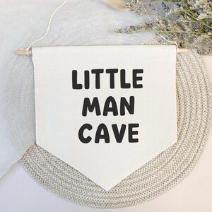 May include: White canvas banner with black text that reads "Little Man Cave". The banner is hanging from a wooden dowel rod and is displayed against a light beige background.