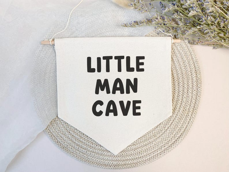 May include: White canvas banner with black text that reads "Little Man Cave". The banner is hanging from a wooden dowel rod and is displayed against a light beige background.