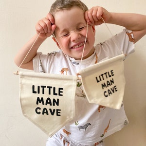 May include: A young child holds up two white fabric banners with black text that reads "Little Man Cave". The child is wearing a white t-shirt with a jungle animal print and white pants.