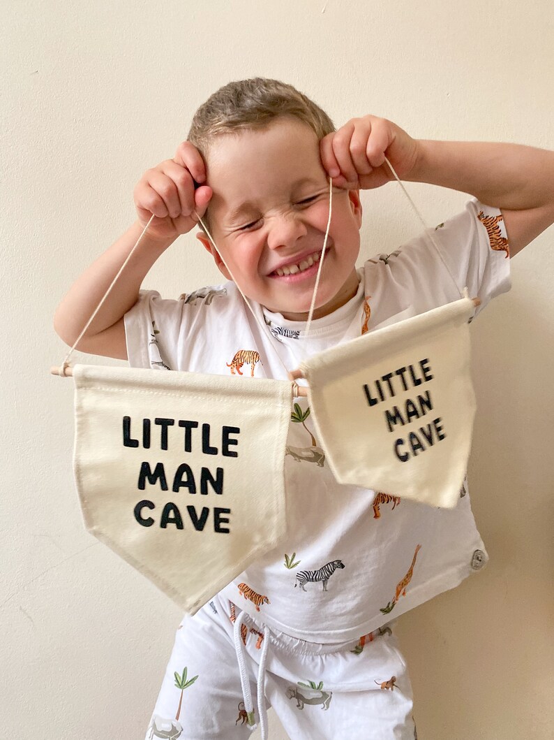 May include: A young child holds up two white fabric banners with black text that reads "Little Man Cave". The child is wearing a white t-shirt with a jungle animal print and white pants.