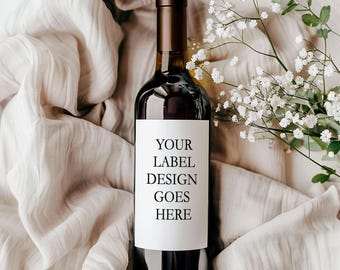 May include: A dark glass wine bottle with a tall neck and a white rectangular label featuring the black text YOUR LABEL DESIGN GOES HERE. The bottle is positioned among soft, light-colored fabric folds and a sprig of small white flowers.
