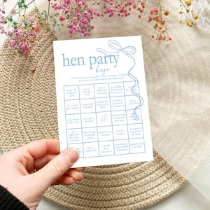 以下が含まれることがあります： 青いリボンデザインの青と白のヘンパーティービンゴカード。カードは5x5のグリッドになっており、各マスにはヘンパーティー中に完了するアクティビティが記載されています。例えば、「見知らぬ人と写真を撮る」「テキーラをショットで飲む」などです。