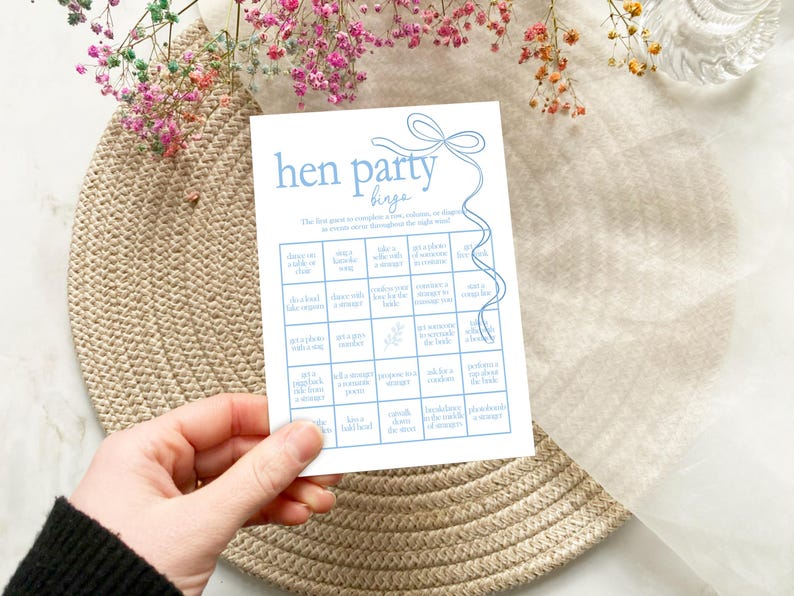 以下が含まれることがあります： 青いリボンデザインの青と白のヘンパーティービンゴカード。カードは5x5のグリッドになっており、各マスにはヘンパーティー中に完了するアクティビティが記載されています。例えば、「見知らぬ人と写真を撮る」「テキーラをショットで飲む」などです。