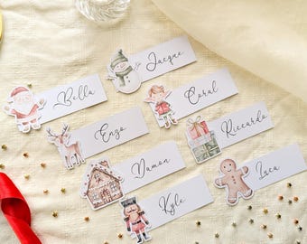 May include: A set of white rectangular name tags featuring watercolor Christmas illustrations including a snowman, Santa, reindeer, gingerbread house, elf, gift boxes, nutcracker, and gingerbread man, with names Bella, Jacque, Enzo, Coral, Damon, Riccardo, Kyle, and Luca.