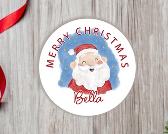 May include: A circular white sticker featuring a watercolor illustration of Santa Claus with a red hat and white beard against a blue background with yellow stars. The text Merry Christmas is printed in red above, and the name Bella is below.