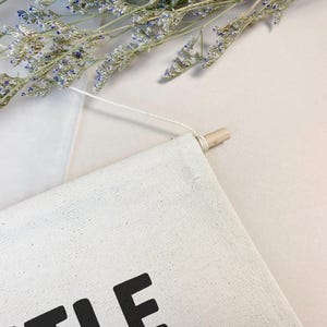 May include: A white canvas wall hanging with the word "Little" printed in black. The hanging is attached to a wooden dowel with white string.