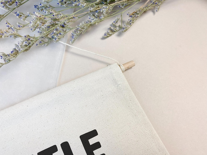 May include: A white canvas wall hanging with the word "Little" printed in black. The hanging is attached to a wooden dowel with white string.