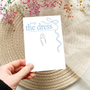以下が含まれることがあります： 青い文字で「ドレスを当てて」と書かれた白いカードと、人の線画が描かれています。カードは青いリボンで飾られています。