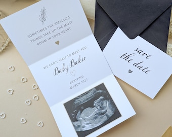 May include: A white folded announcement card featuring a black and white ultrasound image, text reading Sometimes the smallest things take up the most room in your heart, We can't wait to meet you Baby Baker Arriving March 2027, and a save the date card.