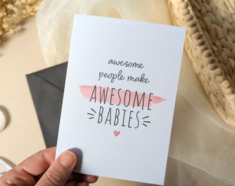 May include: A white greeting card with black text that reads awesome people make AWESOME BABIES. The word AWESOME is highlighted by a horizontal pink brushstroke, and a small pink heart is positioned below the text. The card is rectangular.