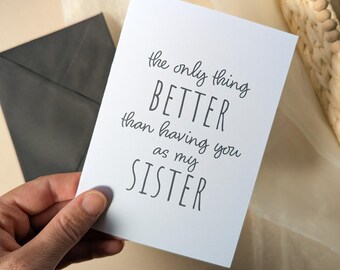 May include: A white greeting card with black printed text that reads: the only thing BETTER than having you as my SISTER. The card is rectangular with a smooth finish and is presented alongside a dark gray envelope.