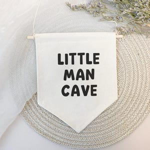 May include: A white canvas banner with black text that reads "Little Man Cave". The banner is hanging from a wooden dowel rod and is displayed against a light beige background.