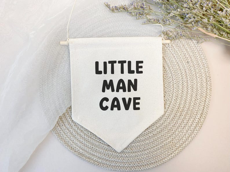 May include: A white canvas banner with black text that reads "Little Man Cave". The banner is hanging from a wooden dowel rod and is displayed against a light beige background.