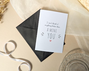 May include: A white greeting card with black text that reads I can't think of anything better than A MINI YOU, featuring a small pink heart below the text, resting on top of a dark charcoal gray envelope with a triangular flap.