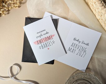 May include: A white greeting card with black text that reads awesome people make AWESOME BABIES with a pink brush stroke and a small heart. Next to it is an open card with the text Baby Smith ARRIVING MAY 2027. A black envelope is underneath.