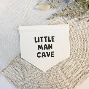 May include: A white canvas banner with the text "LITTLE MAN CAVE" in black lettering. The banner is hanging from a wooden dowel and is displayed against a light brown background.