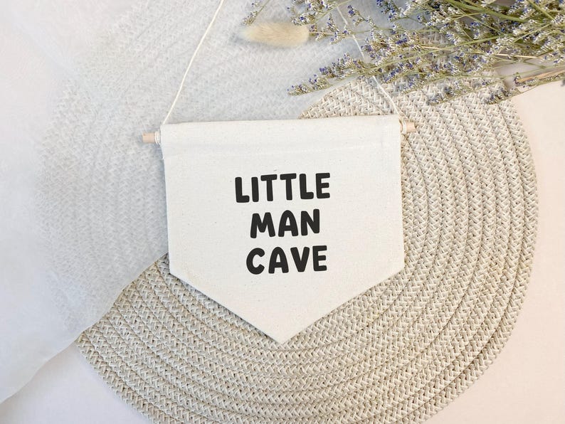 May include: A white canvas banner with the text "LITTLE MAN CAVE" in black lettering. The banner is hanging from a wooden dowel and is displayed against a light brown background.