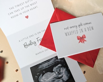 May include: A white folded announcement card featuring a black and white ultrasound image, red envelopes, and text that reads: THE TINIEST GIFTS CAN TAKE UP THE MOST ROOM IN YOUR HEART, A LITTLE ONE IS ON THE, Baby, MARCH 2027, and not every gift comes WRAPPED IN A BOW.