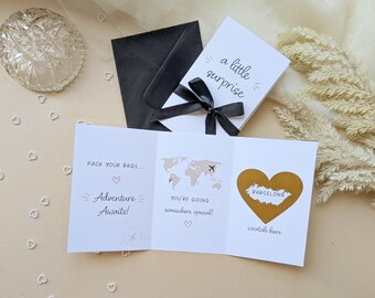 May include: A white tri-fold card with black text reading "a little surprise" on the front, tied with a black ribbon. Inside, the text reads "PACK YOUR BAGS... Adventure Awaits!", "YOU'RE GOING somewhere special!" with a world map, and a gold heart with "BARCELONA" and "scratch here" printed below it, accompanied by a black envelope.