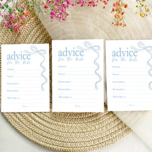 以下が含まれることがあります： 青いリボン模様の白いカードが3枚あり、それぞれに「花嫁へのアドバイス」という文字が印刷されています。各カードには、ゲストが花嫁へのアドバイスを書くためのプロンプトがリストされており、「常に」、「決して」、「時々」、「覚えておいて」、「結婚とは」などが含まれています。