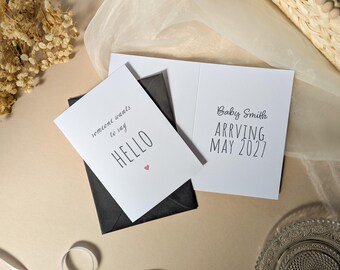 May include: A white folded greeting card with the text "someone wants to say HELLO" and a small red heart on the front, paired with a black envelope. The inside of the card displays the text "Baby Smith ARRIVING MAY 2027" in black lettering.